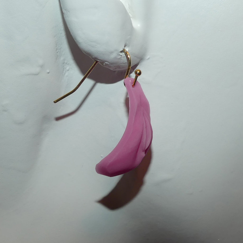 A single pink petal-shaped vulva dangle earring with a gold hook, shown hanging against a white textured wall.