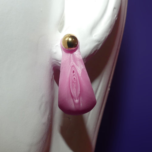 A pink silicone novelty stud earring shaped like a stylized vulva, featuring a gold-tone ball stud at the top, displayed on a white clay ear stand against a dark purple background.