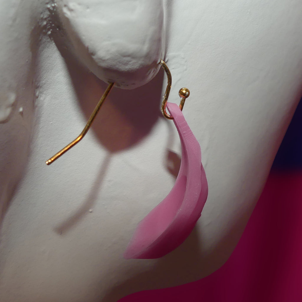 A close-up of a pink petal-shaped barbie vagina dangle earring with a gold hook, displayed on a white mannequin bust against a pink background.