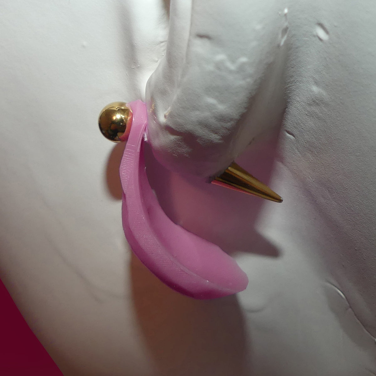 A close-up of a pink curved barbie vagina earring with a gold-tone ball and spike, shown on a white mannequin ear.