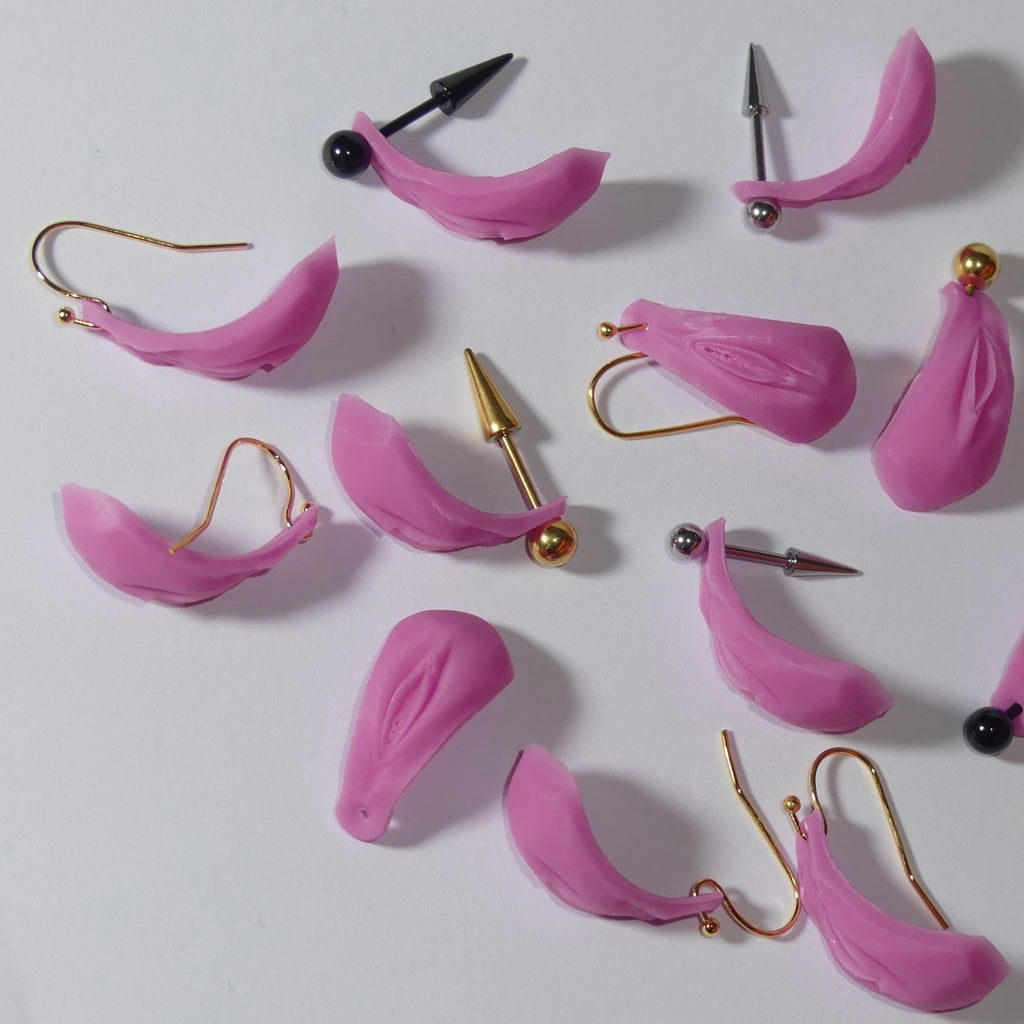 A collection of pink silicone vulva Vagina ear cuffs with various metal ear hooks, including gold, silver, and black options, arranged on a plain white surface.