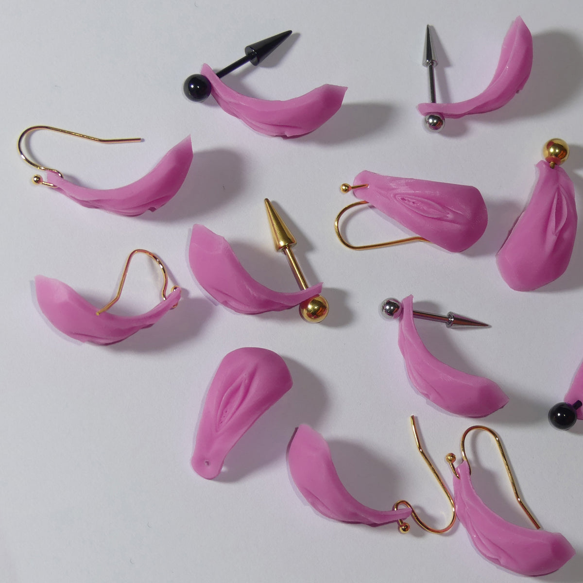 A collection of pink silicone vulva Vagina ear cuffs with various metal ear hooks, including gold, silver, and black options, arranged on a plain white surface.