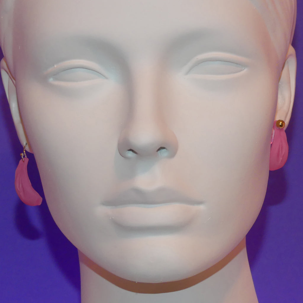 Pink crescent-shaped vulva dangle earrings displayed on a white mannequin head against a purple background.