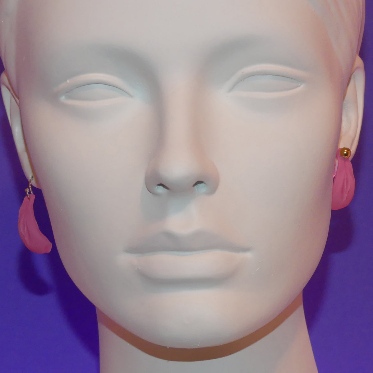 Pink crescent-shaped vulva dangle earrings displayed on a white mannequin head against a purple background.