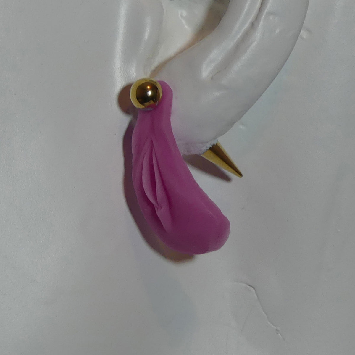 A pink vagina dangle earring with a gold ball stud and gold spike back, shown on a white ear form a white mannequin.