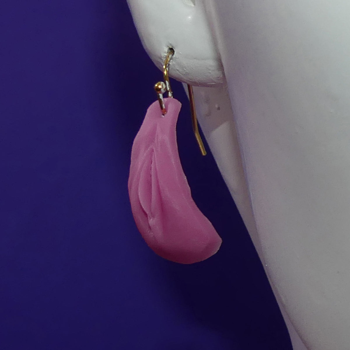 A single pink vulva teardrop-shaped drop earring with a gold fishhook clasp, shown hanging from a white mannequin ear against a solid dark purple background.