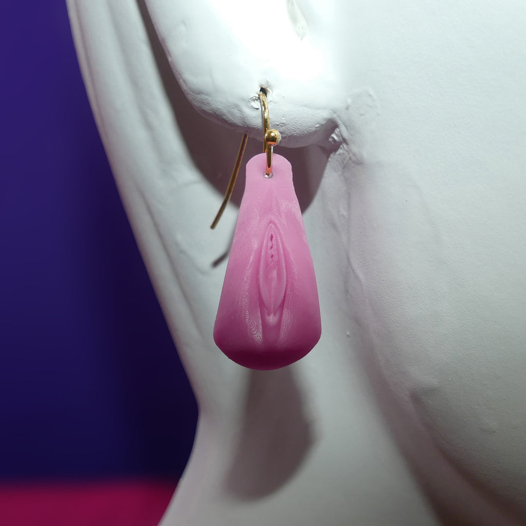 A pink vulva Vagina -shaped dangle earring with a gold fishhook wire, displayed on a white female mannequin bust against a purple and pink background.