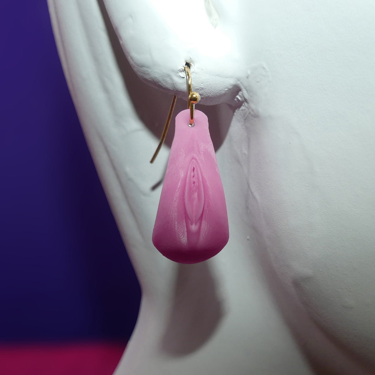 A pink vulva Vagina -shaped dangle earring with a gold fishhook wire, displayed on a white female mannequin bust against a purple and pink background.