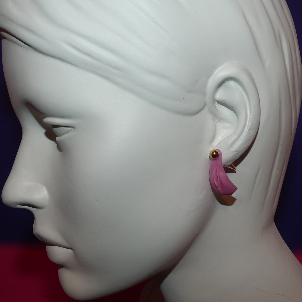 A pink vulva and gold ball and spike earring displayed on the ear of a white mannequin head in profile against a dark blue and red background.