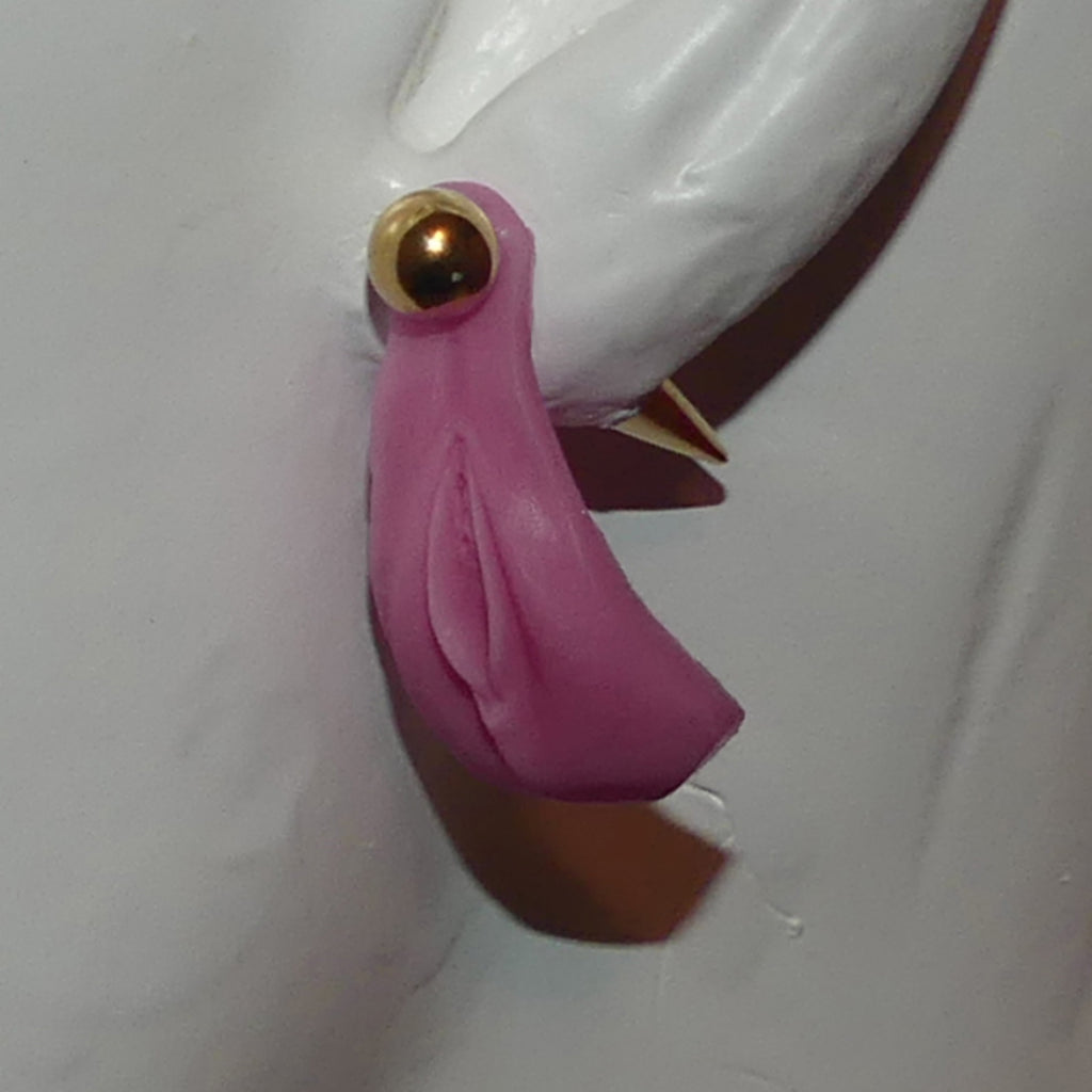A close-up of a pink sculpted vulva with a gold-toned circular stud and a small pointed gold accent, attached to a white mannequin form against a light gray background.