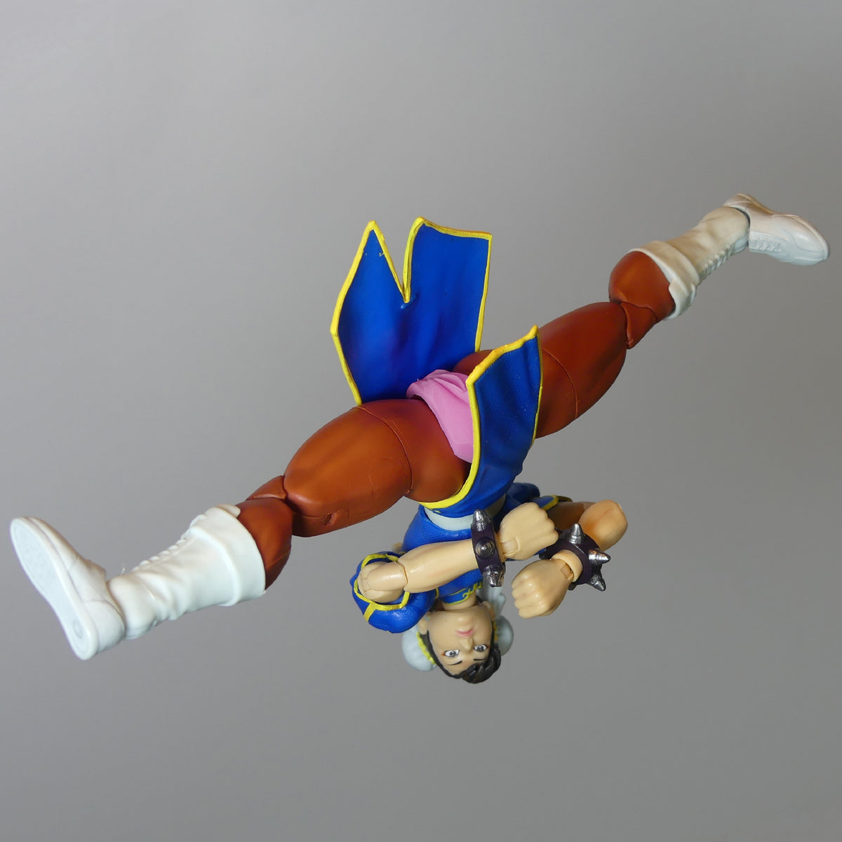 A Street Fighter figurine of Chun-Li shown upside down in a flying pose, wearing her signature blue and pink outfit with brown pants and white boots, set against a plain gray background.