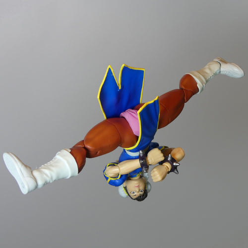 A Street Fighter figurine of Chun-Li shown upside down in a flying pose, wearing her signature blue and pink outfit with brown pants and white boots, set against a plain gray background.