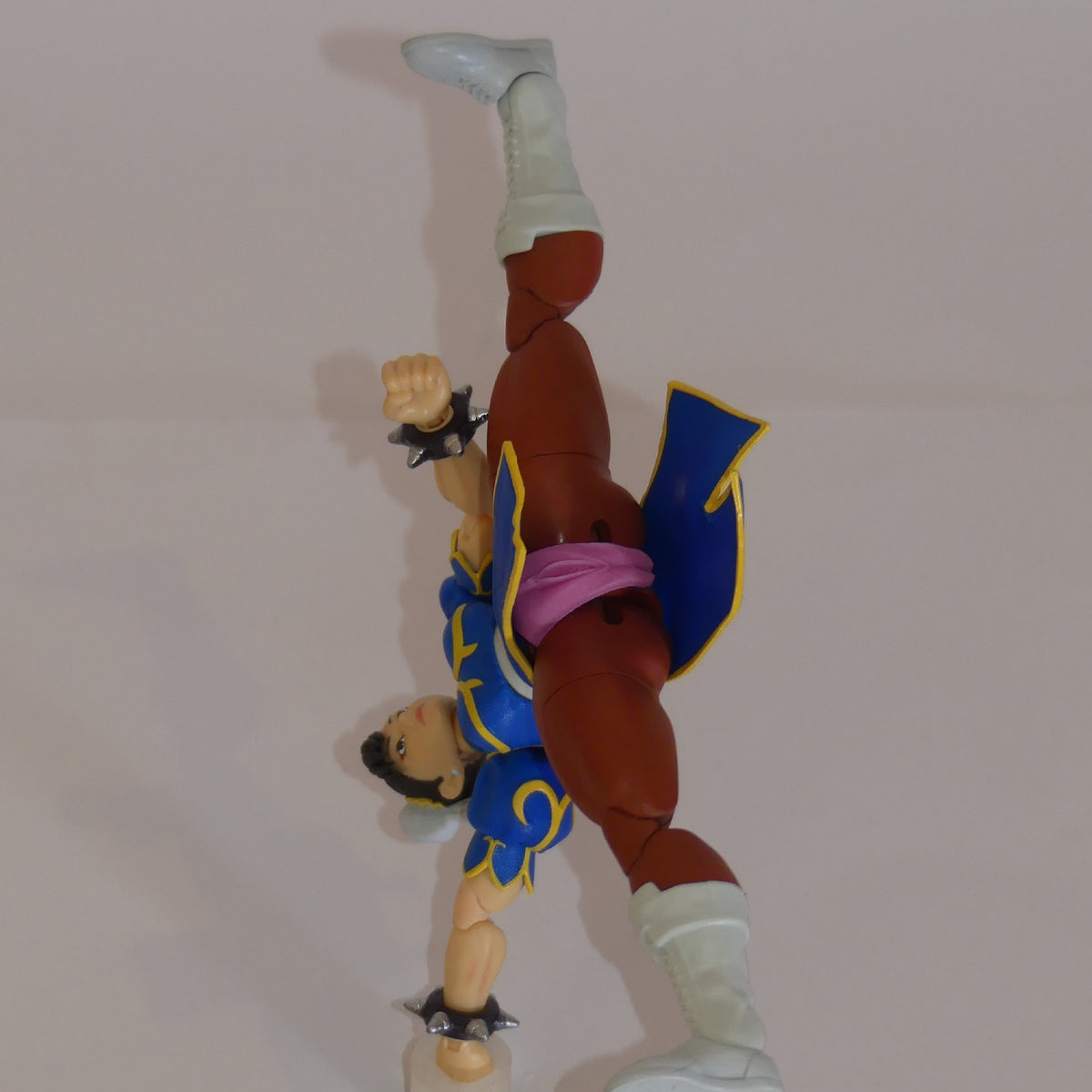 A Street Fighter 5 action figure of Chun-Li posed dynamically in a handstand, wearing her signature blue and gold outfit with a pink vagina, white boots, and brown skin.